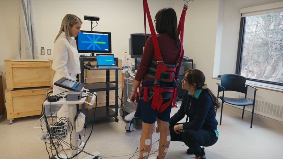 Person wearing a safety harness participating in a balance rehabilitation assessment while researchers monitor equipment in a laboratory setting.